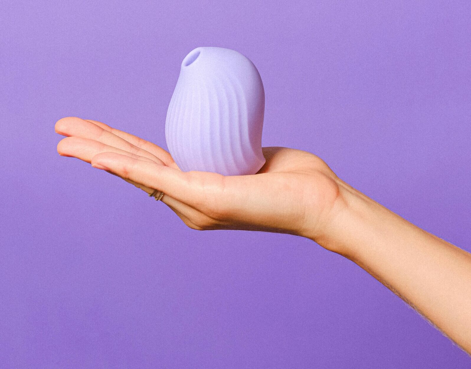 Succionador de clítoris de silicona violeta sostenido en una mano, fondo lila